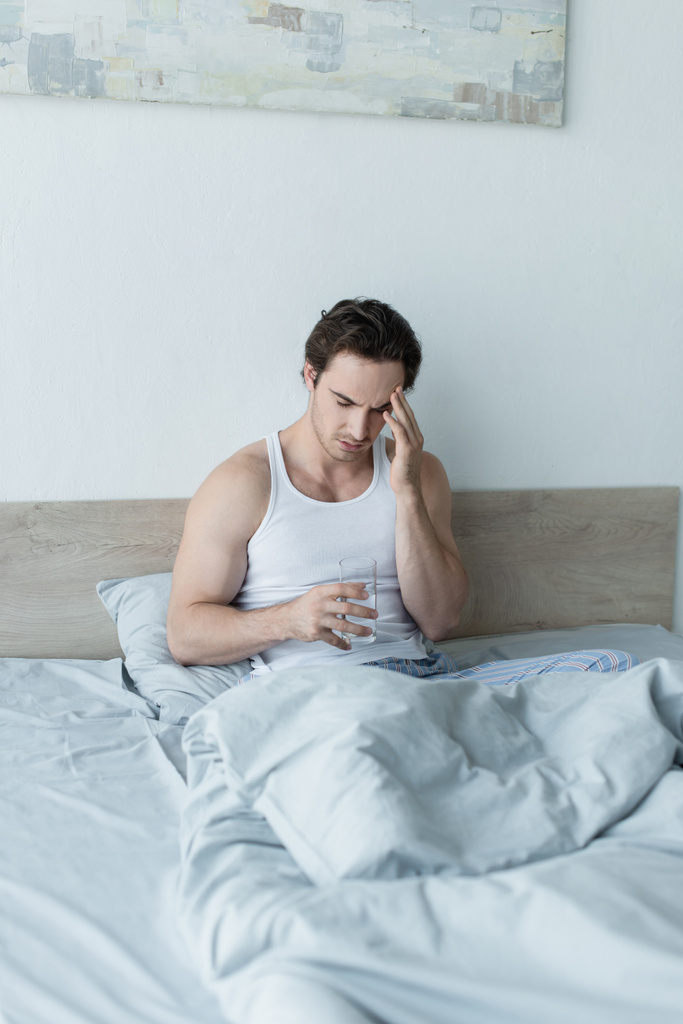 Junger Mann sitzt mit Glas Wasser im Bett und leidet unter Kopfschmerzen - Foto, Bild