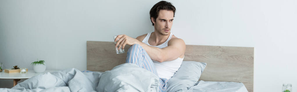 καταθλιπτικός άντρας κοιτάζει αλλού ενώ κάθεται στο κρεβάτι με ένα ποτήρι νερό, πανό - Φωτογραφία, εικόνα