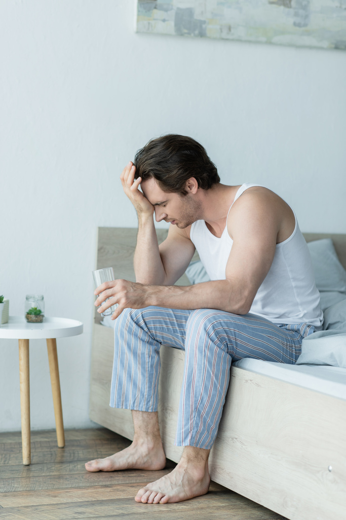 mężczyzna w piżamie cierpiący na ból głowy siedząc na łóżku ze szklanką wody - Zdjęcie, obraz