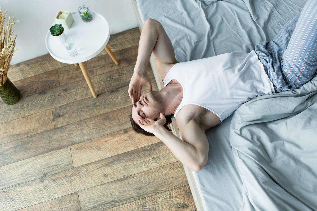 high angle view of man lying on edge of bed while suffering from hangover in morning - Photo, Image