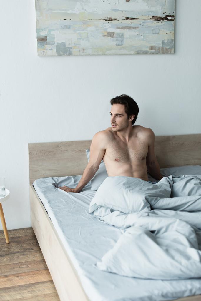 Hemdloser Mann sitzt unter blauer Decke im Bett und schaut weg - Foto, Bild