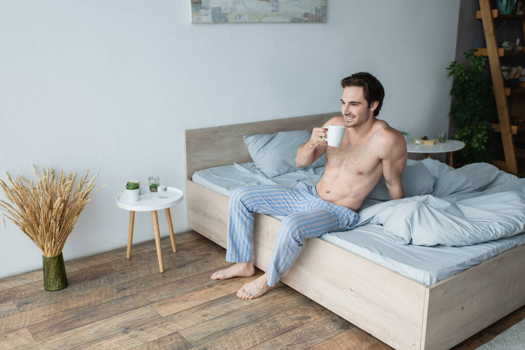 ベッドルームでコーヒーを飲みながら微笑む縞模様のパジャマパンツの白人男性 - 写真・画像