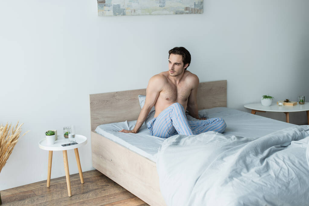 giovane uomo senza maglietta seduto sul letto al mattino e guardando altrove - Foto, immagini