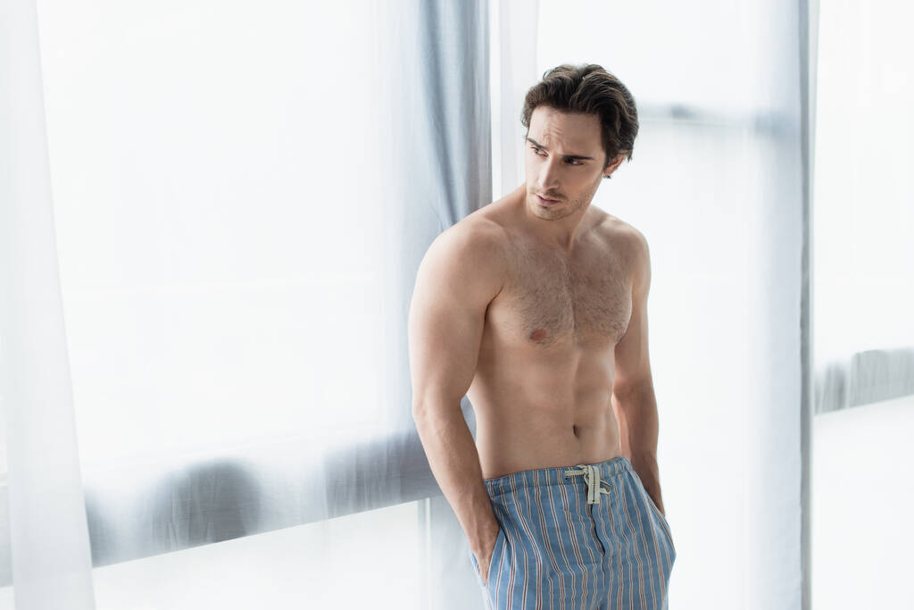 homme torse nu debout près de la fenêtre avec les mains dans des poches de pantalon de pyjama - Photo, image