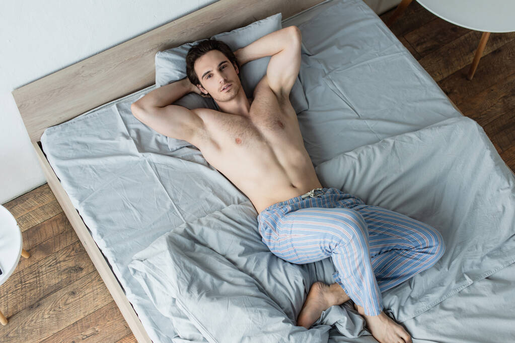 hoge hoek uitzicht van shirtloze man liggend in bed met handen achter het hoofd - Foto, afbeelding