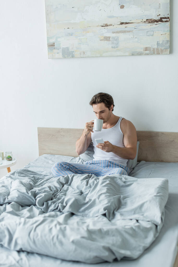 νεαρός άνδρας που κάθεται στο κρεβάτι, πίνοντας καφέ και ανταλλάσσοντας μηνύματα στο κινητό τηλέφωνο - Φωτογραφία, εικόνα