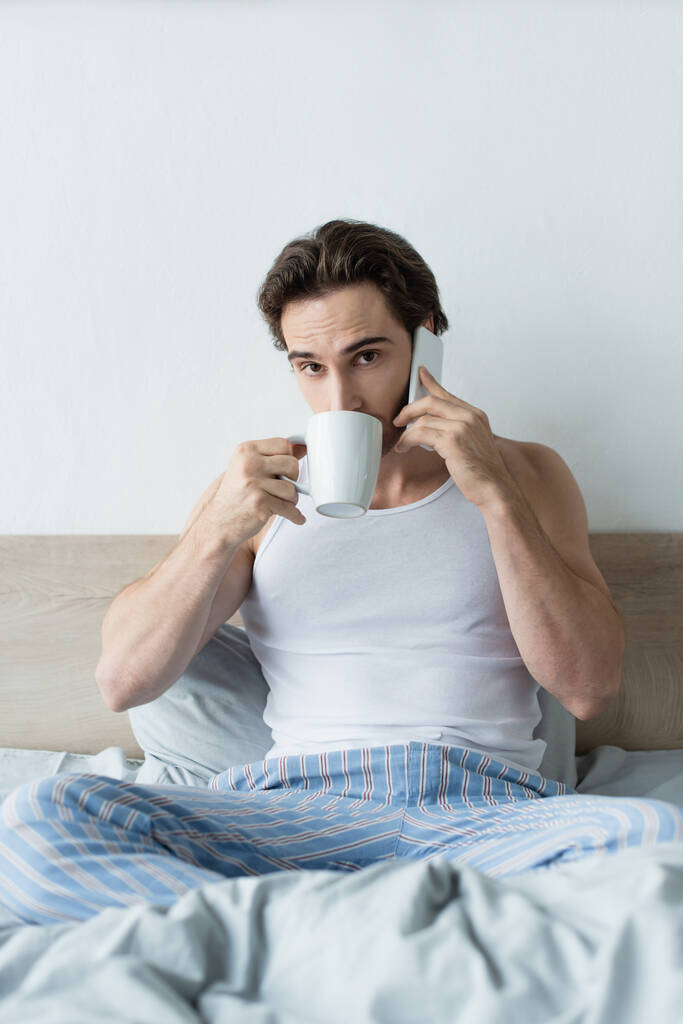 νεαρός άνδρας πίνει καφέ και μιλάει στο κινητό ενώ κάθεται στο κρεβάτι - Φωτογραφία, εικόνα