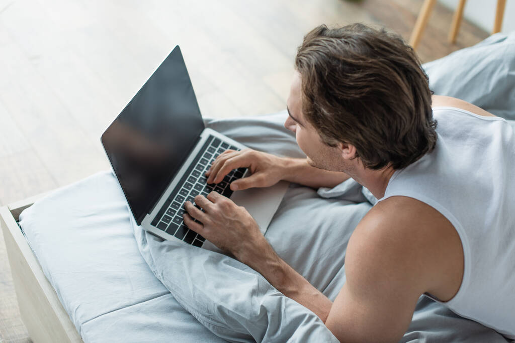 високий кут зору брюнетки чоловік друкує на ноутбуці з порожнім екраном в ліжку
 - Фото, зображення