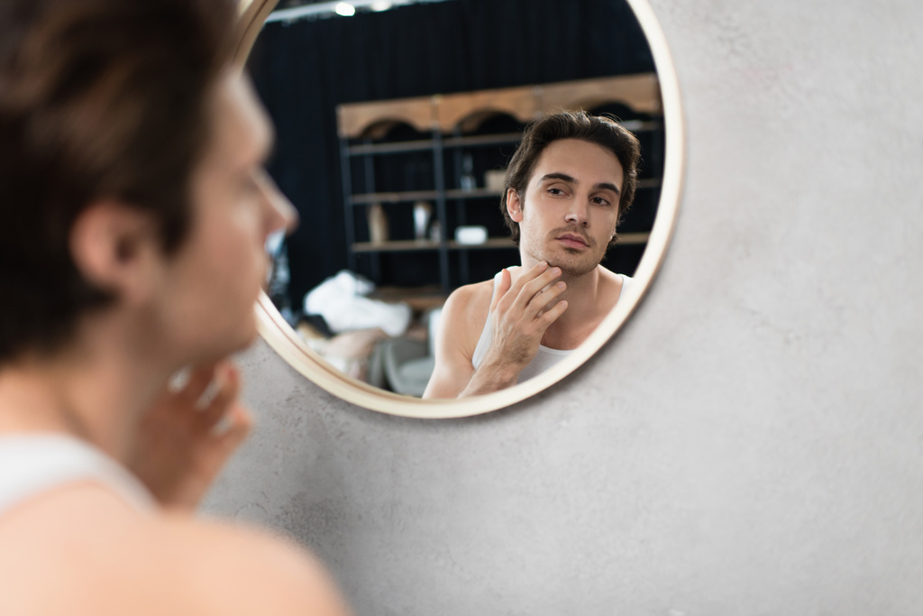 jovem homem tocando rosto raspado enquanto olha no espelho - Foto, Imagem
