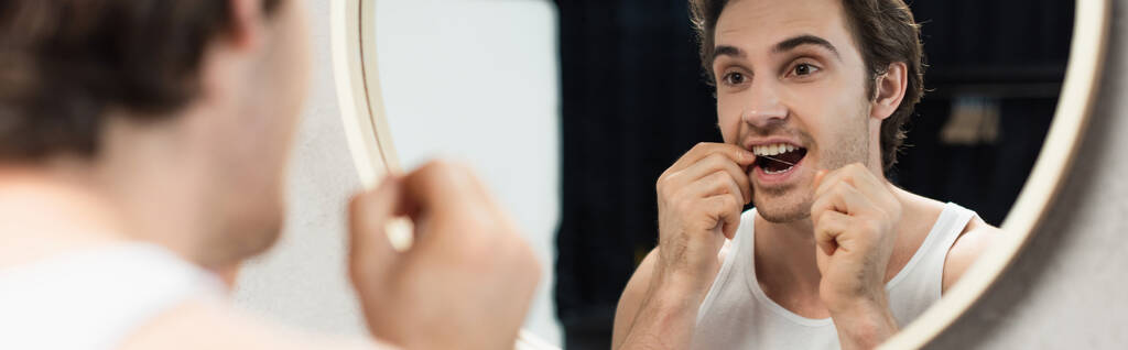 młody człowiek patrząc w lustro podczas nici dentystycznej zębów rano, baner - Zdjęcie, obraz