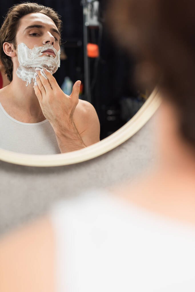 fiatal férfi alkalmazása borotválkozás hab az arcon tükör közelében a fürdőszobában elmosódott előtérben - Fotó, kép