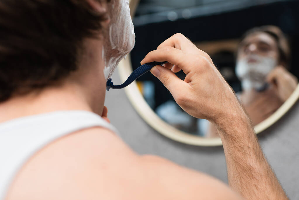 размытый человек бритья с безопасной бритвой возле зеркала в ванной комнате - Фото, изображение
