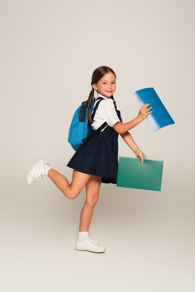 volledige weergave van vrolijke schoolkind lopen met notebooks op grijs - Foto, afbeelding