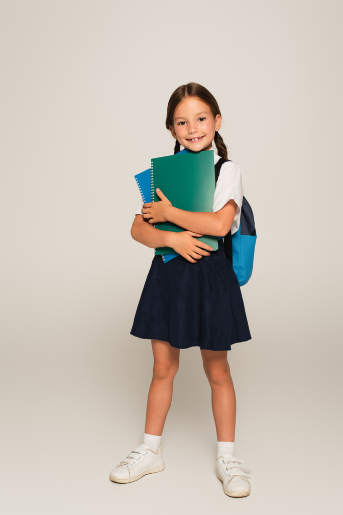 full length view of joyful schoolgirl standing with copy books on grey - Photo, Image