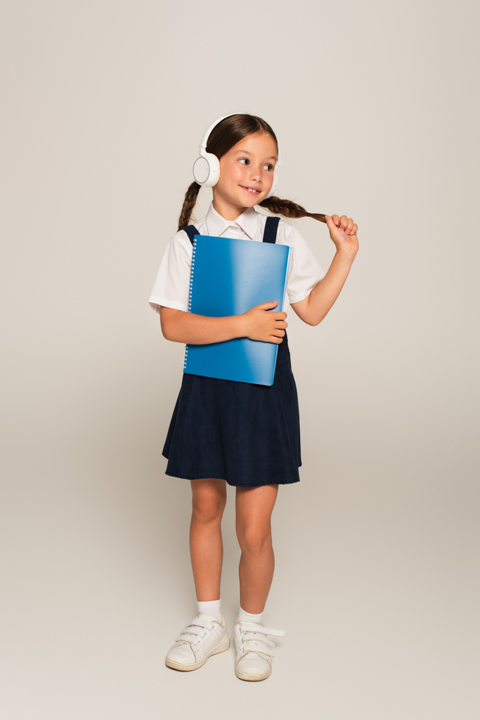 pozitivní školačka ve sluchátkách dotýkající se copytail při držení kopie knihy na šedé - Fotografie, Obrázek