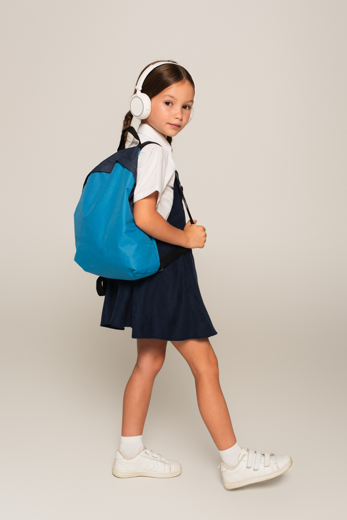 Schülerin mit Kopfhörer läuft mit blauem Rucksack und blickt in Kamera auf grau - Foto, Bild