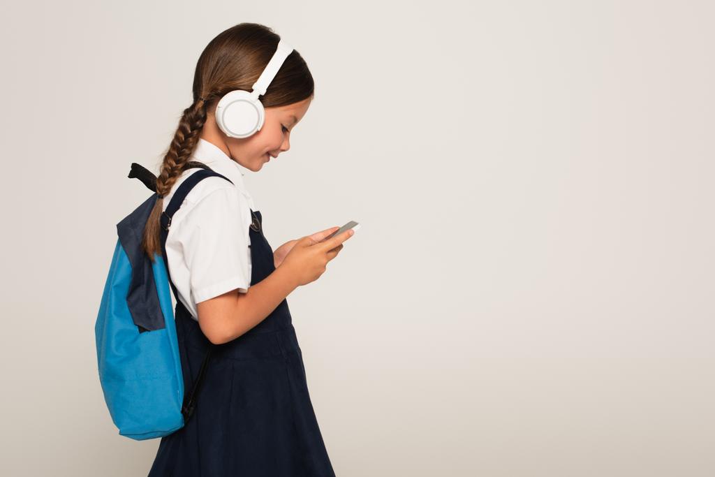 boční pohled na veselý školák ve sluchátkách pomocí smartphone izolované na šedé - Fotografie, Obrázek