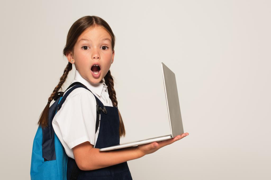 κατάπληκτη μαθήτρια με ανοιχτό στόμα κοιτάζοντας κάμερα, ενώ κρατώντας φορητό υπολογιστή απομονωμένο σε γκρι - Φωτογραφία, εικόνα