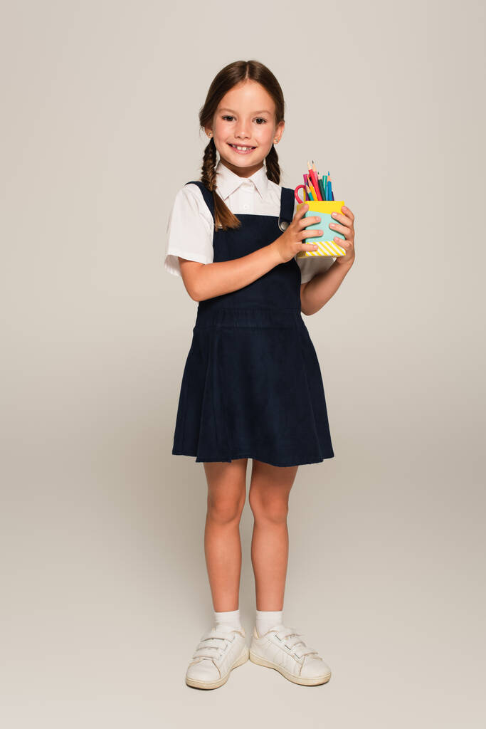 fröhliches Schulmädchen hält Stifthalter mit Bleistiften und Filzstiften auf grau - Foto, Bild