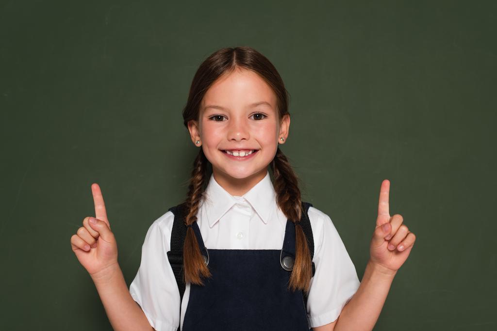 χαρούμενο κορίτσι με σχολική στολή δείχνει με τα δάχτυλα κοντά σε μαυροπίνακα, ενώ κοιτάζοντας την κάμερα - Φωτογραφία, εικόνα