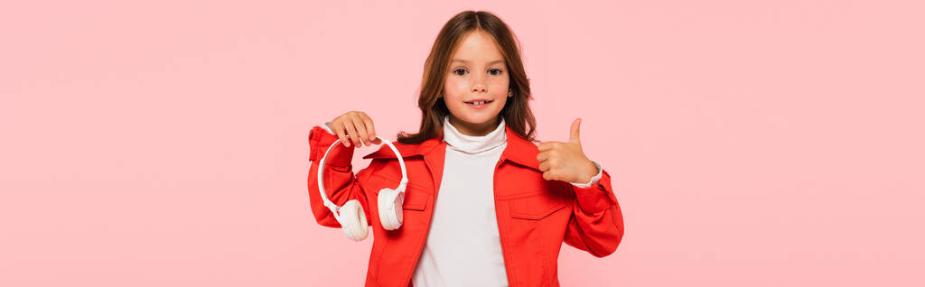 elegante chica sosteniendo los auriculares y mostrando el pulgar hacia arriba aislado en rosa, bandera - Foto, Imagen