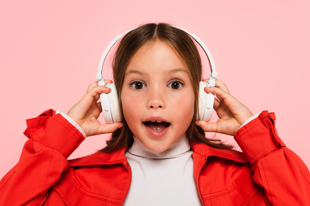 amazed girl with open mouth adjusting headphones isolated on pink - Photo, Image
