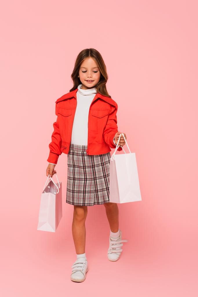 meisje op oranje jas en geruite rok lopen met boodschappentassen op roze - Foto, afbeelding