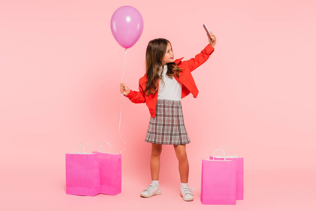 trendy kid taking selfie on smartphone while standing with balloon near shopping bags on pink - Photo, Image