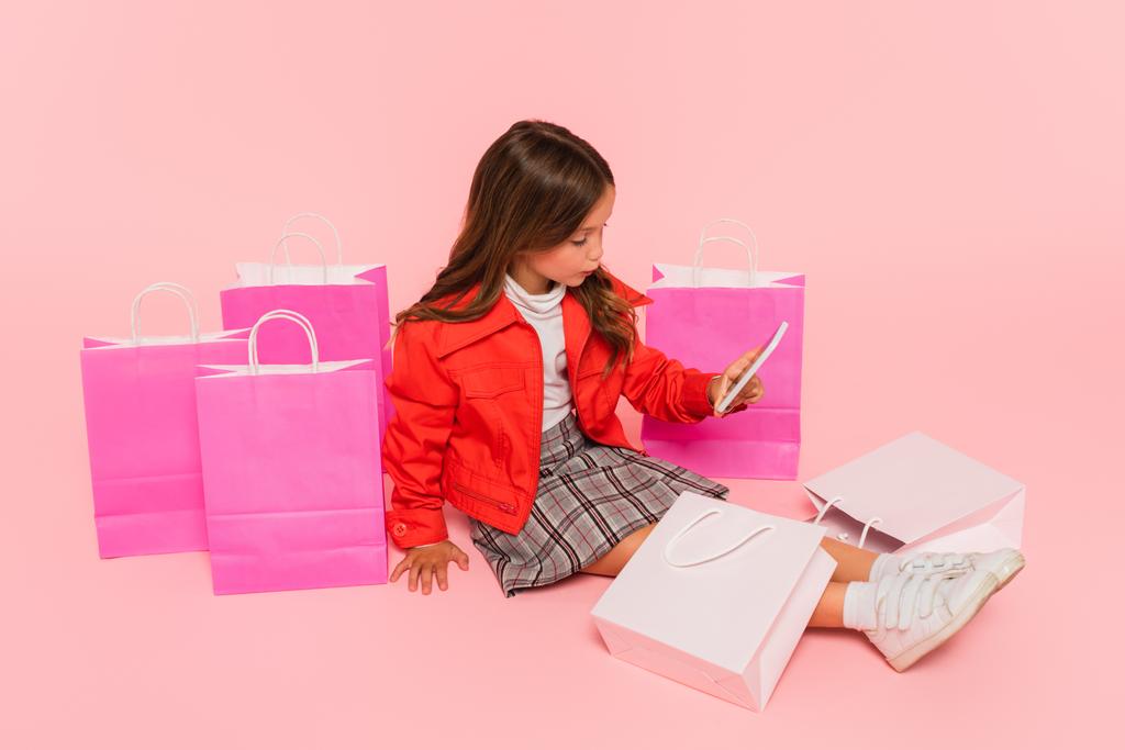 流行の服を着た女の子がピンクのショッピングバッグの近くに座って - 写真・画像