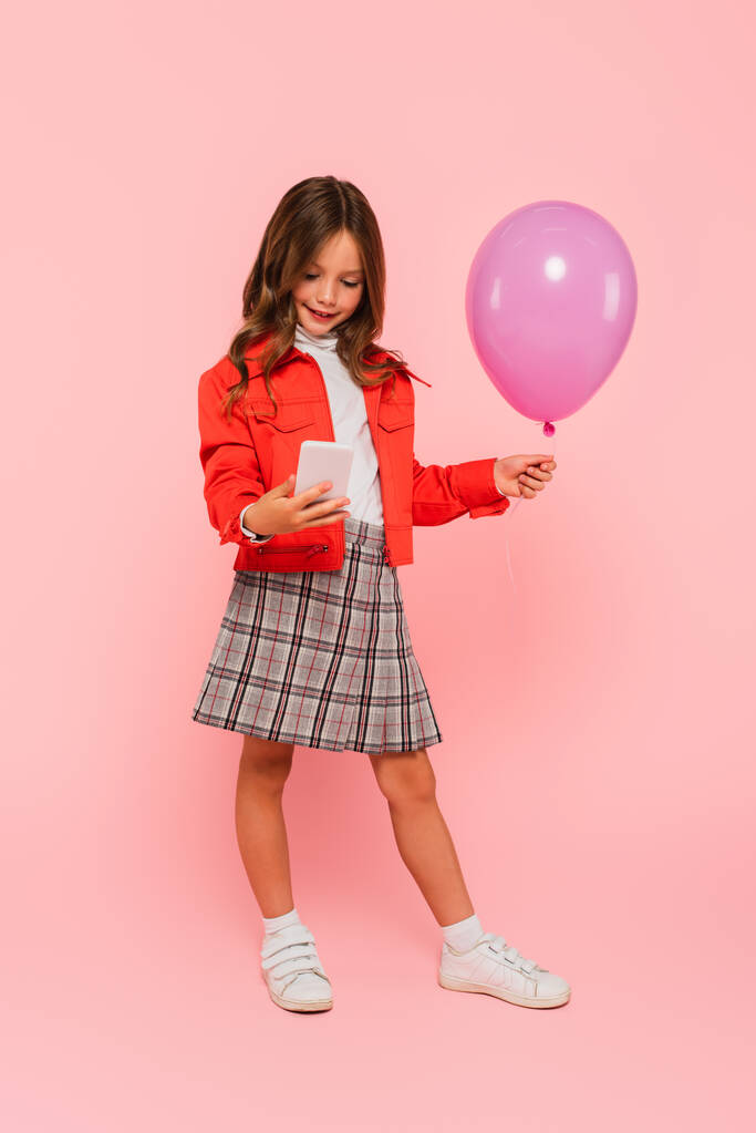 παιδί σε μοντέρνα ρούχα στέκεται με κινητό τηλέφωνο και μπαλόνι σε ροζ - Φωτογραφία, εικόνα
