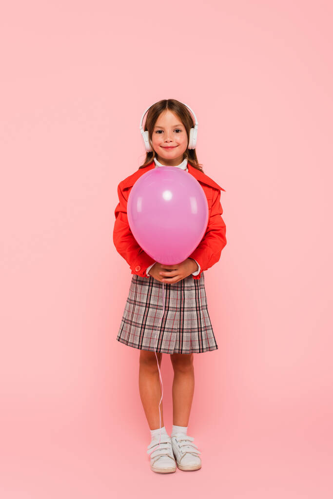 full length view of fashionable child in headphones standing with festive balloon on pink  - Photo, Image