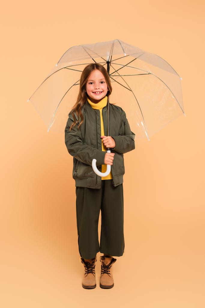 full length άποψη του χαμογελώντας κορίτσι στο σακάκι βομβαρδιστικό στέκεται κάτω από ομπρέλα σε μπεζ - Φωτογραφία, εικόνα