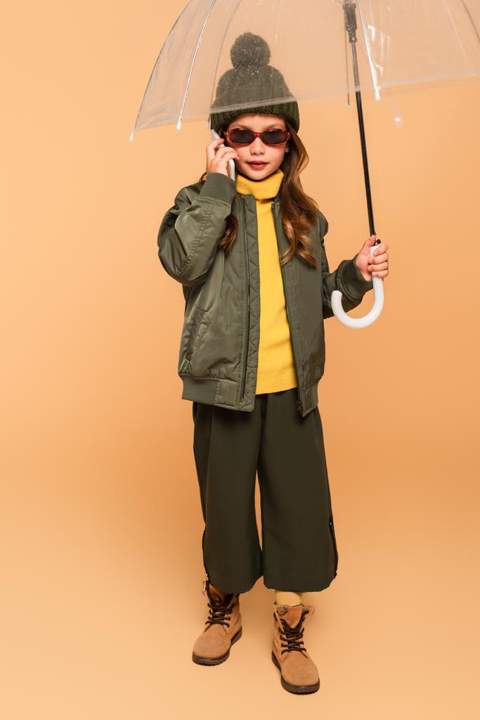 vista completa de la muchacha en ropa de moda hablando en el teléfono celular bajo paraguas en beige - Foto, imagen