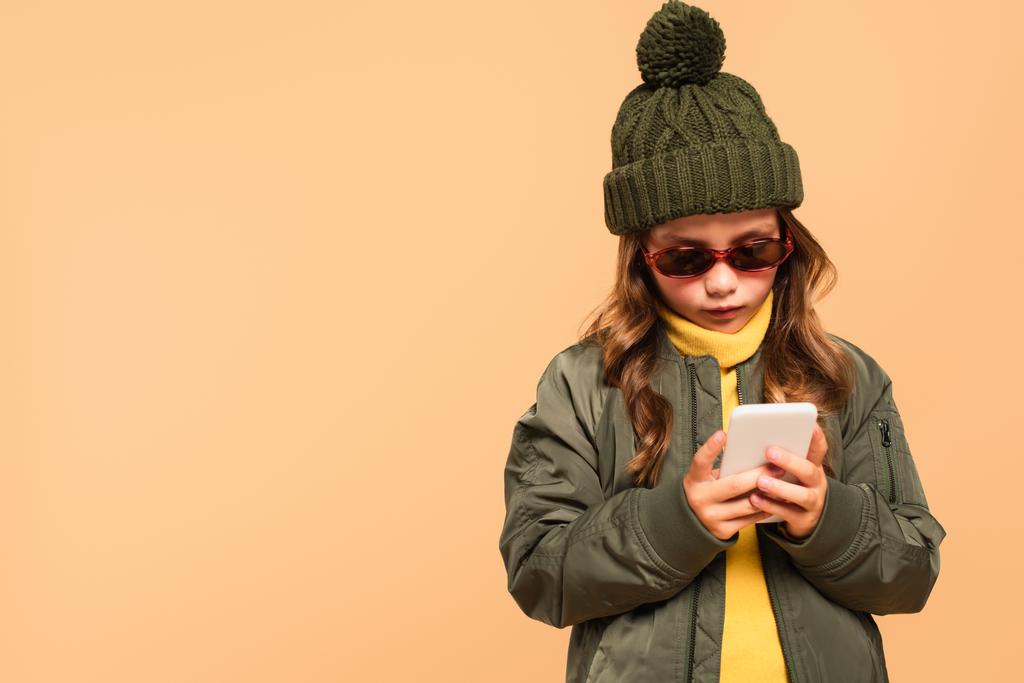 girl in trendy autumn outfit and sunglasses using smartphone isolated on beige - Photo, Image