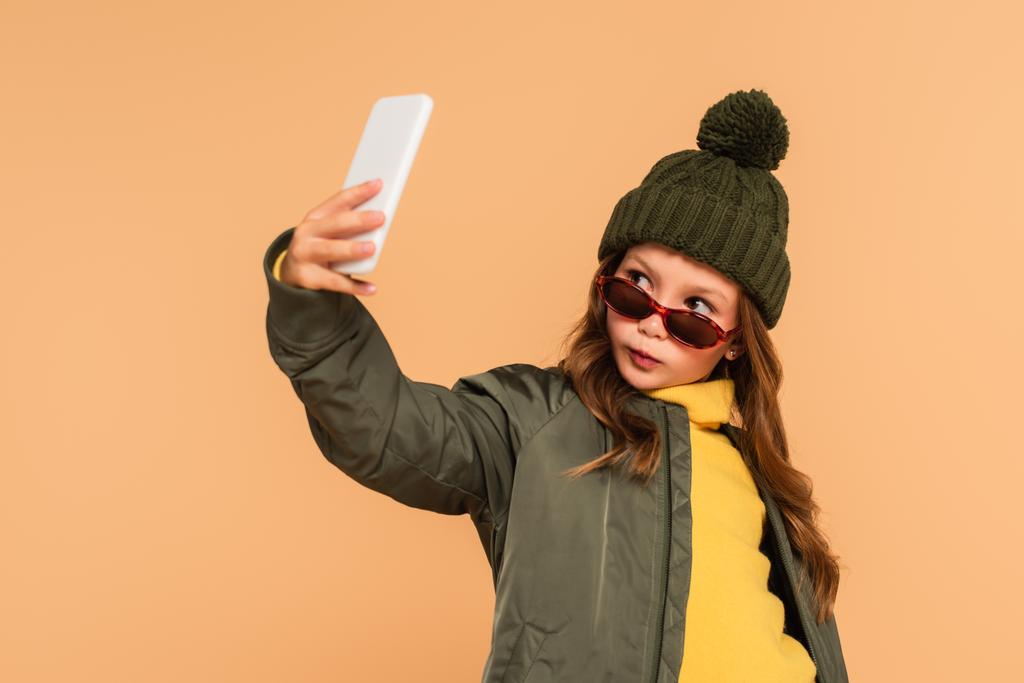 niño con estilo en la ropa de otoño tomar selfie en el teléfono inteligente aislado en beige - Foto, imagen