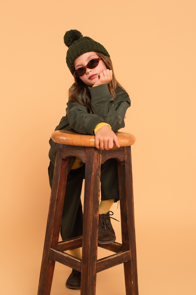 criança em óculos de sol e roupas de outono na moda olhando para a câmera perto de banquinho de madeira no bege - Foto, Imagem