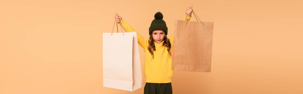 modne dziecko w żółtym golfie trzymające torby na zakupy w podniesionych rękach odizolowane na beżu, sztandar - Zdjęcie, obraz