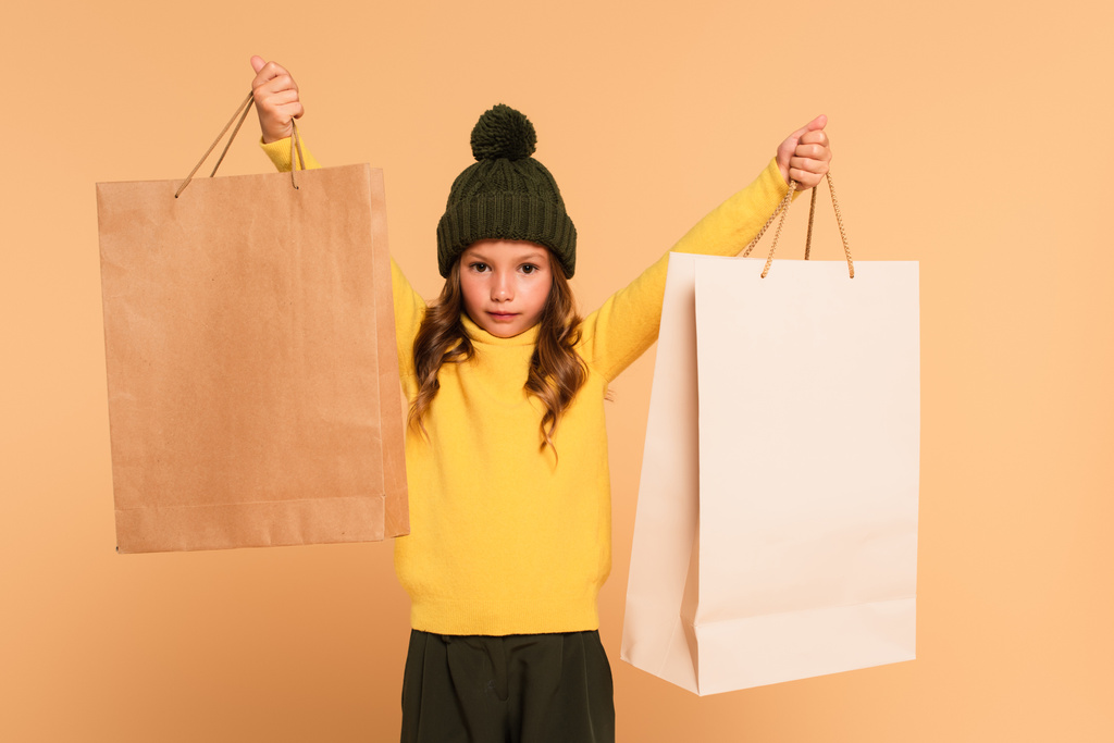 menina na moda em gola alta amarela em pé com sacos de compras em mãos levantadas isolado no bege - Foto, Imagem