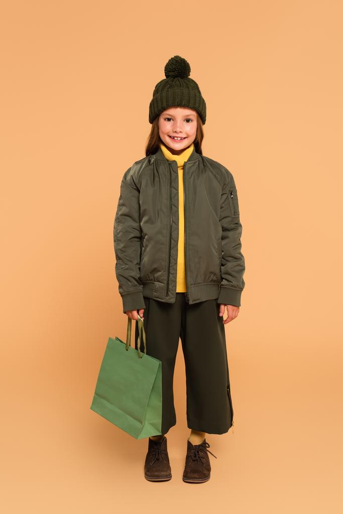 chica feliz en chaqueta de bombardero y sombrero marrón sosteniendo bolsa de compras en beige - Foto, imagen