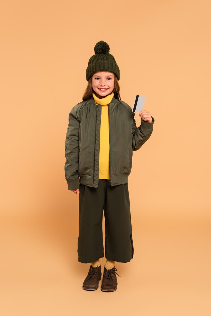 Вид в полный рост счастливой девушки в стильной осенней одежде с кредитной картой на бежевом фоне - Фото, изображение
