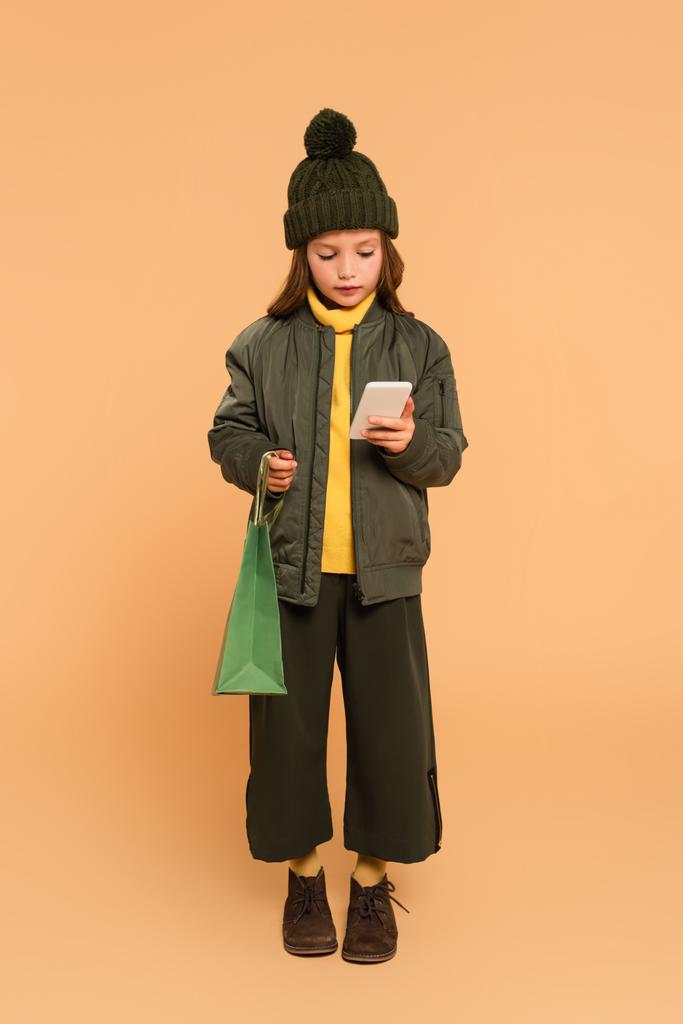 volledige weergave van meisje in trendy herfst outfit met boodschappentas en mobiele telefoon op beige - Foto, afbeelding