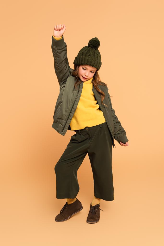full length view of child in fashionable autumn outfit posing with raised hand on beige - Photo, Image
