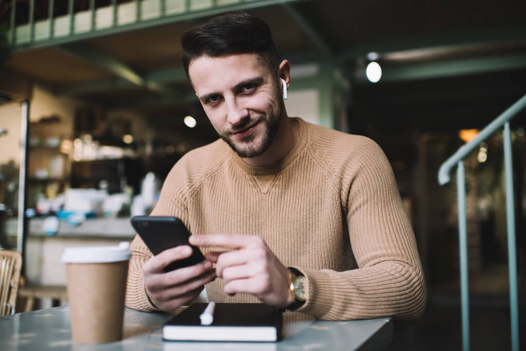 Youthful bearded male in casual wear with wireless earbuds smiling lightly while sitting in cafe using smartphone and looking at camera - Photo, Image