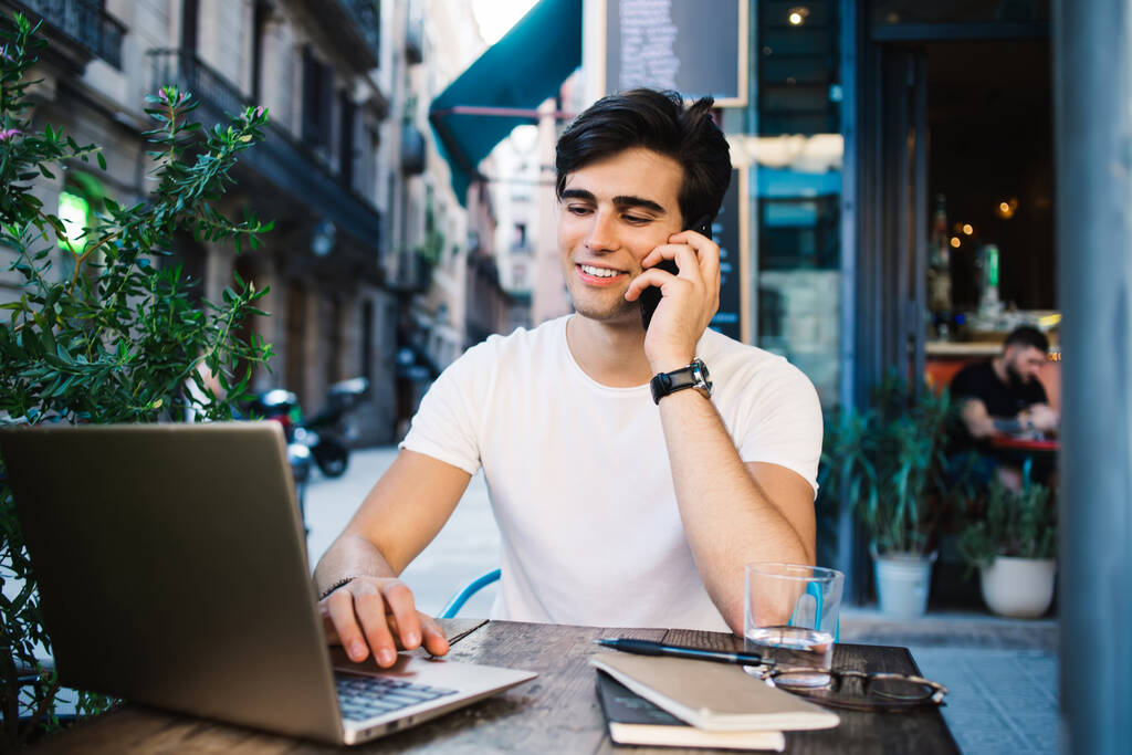 Pozitivní muž pomocí notebooku a mluvit na smartphone při posezení v kavárně s poznámkovými bloky sklenice vody ang brýle na stole - Fotografie, Obrázek