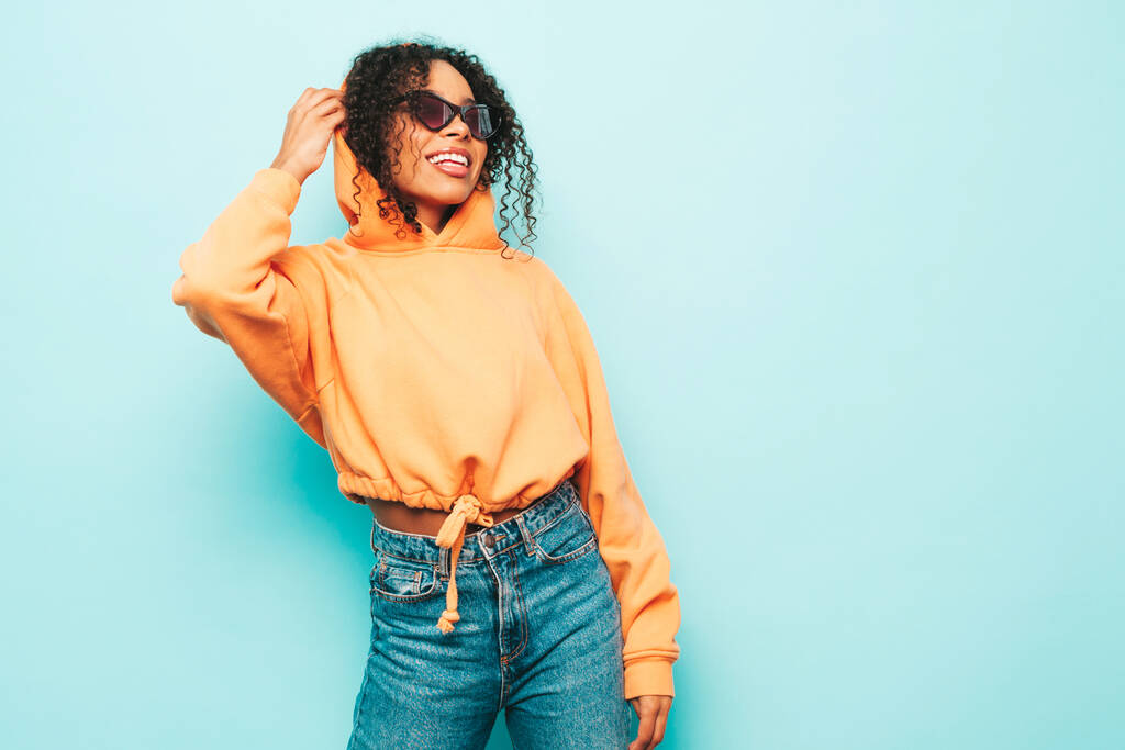 Beautiful black woman with afro curls hairstyle.Smiling model in orange hoodie and trendy jeans clothes. Sexy carefree female posing near blue wall in studio in sunglasses. Cheerful and happy - Photo, Image