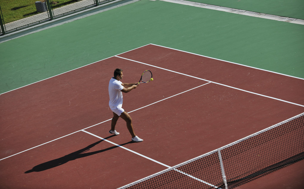 Молодая женщина играет в теннис на открытом воздухе на оранжевом теннисном поле рано утром
 - Фото, изображение