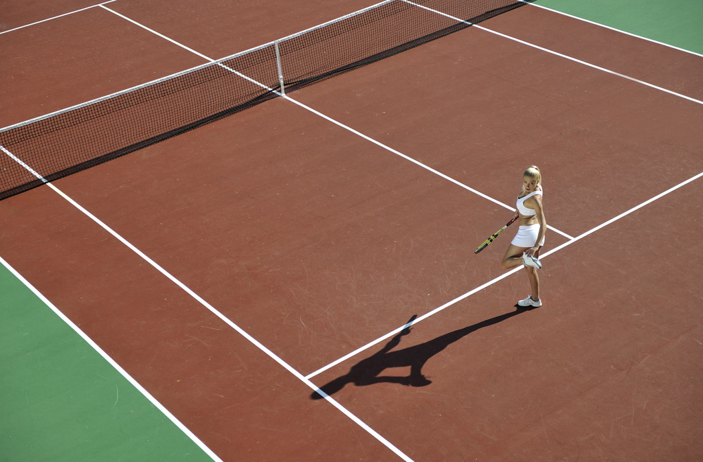 Молодая женщина играет в теннис на открытом воздухе на оранжевом теннисном поле рано утром
 - Фото, изображение