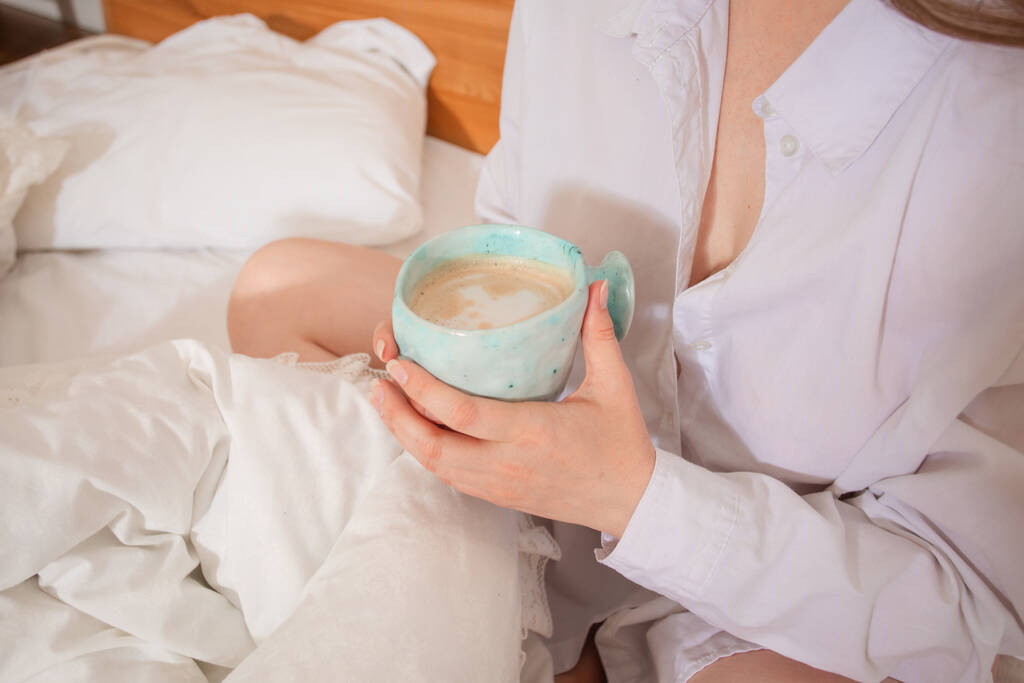 Mano femminile che tiene una tazza di caffè mentre è seduta a letto - Foto, immagini