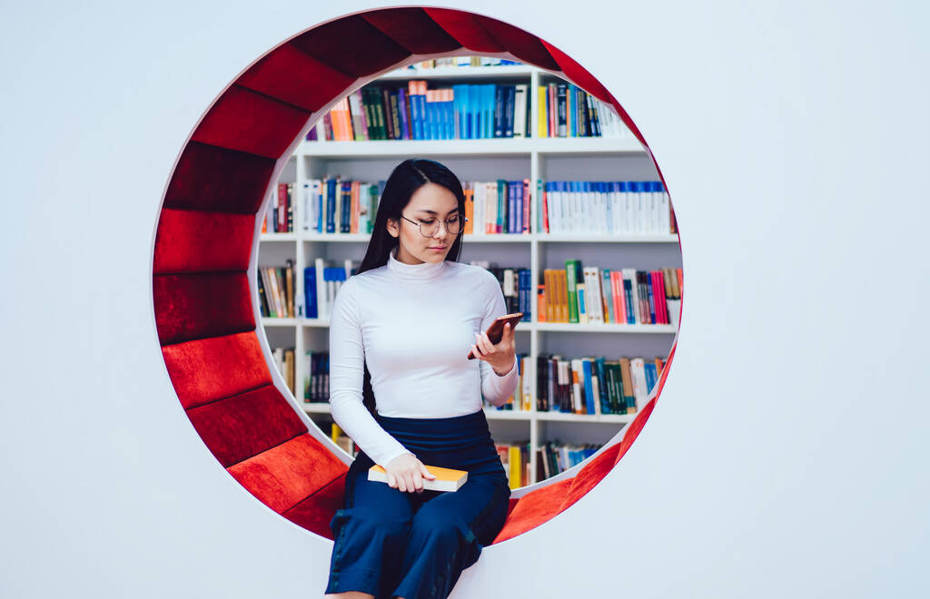 Συγκεντρωμένη εθνοτική γυναίκα σε casual φθορά και γυαλιά κάθεται σε κομψό πάγκο στη σύγχρονη βιβλιοθήκη με βιβλίο, ενώ χρησιμοποιώντας smartphone - Φωτογραφία, εικόνα