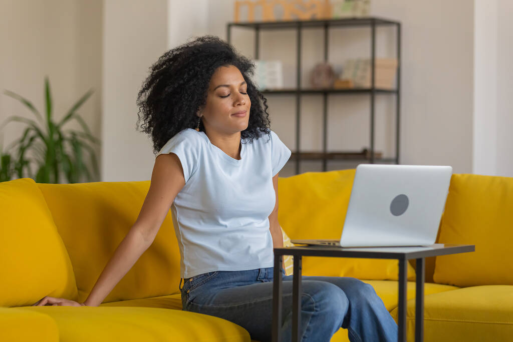 Portrait d'une jeune afro-américaine fatiguée assise les yeux fermés près de l'ordinateur portable. Brunette avec des cheveux bouclés posant sur un canapé jaune dans une pièce de la maison lumineuse. Gros plan. - Photo, image